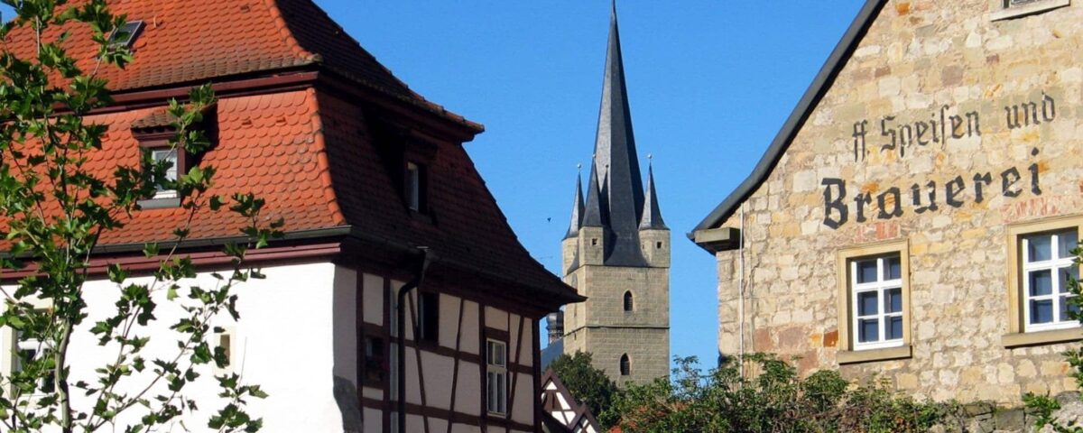 Blick bei schönem Wetter auf eine Kirchturmspitze und eine alte Brauerei im Landkreis Hassberge